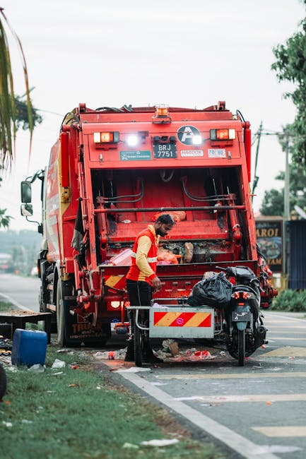 A sanitation worker wearing a high-visibility yellow and red uniform is emptying a large red waste collection truck on a roadside. The truck's rear compartment is open, revealing mechanical components