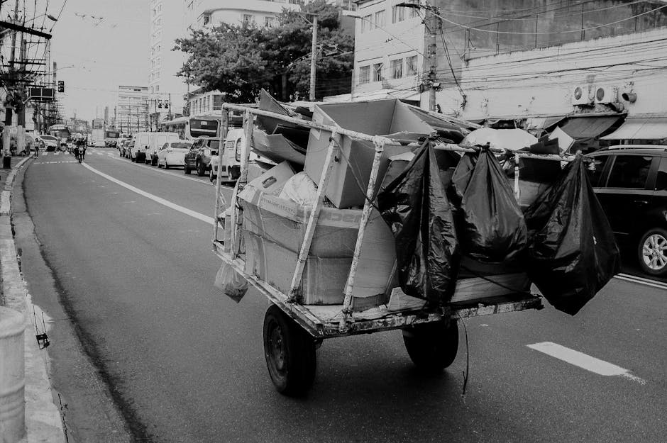 A sanitation worker wearing a high-visibility yellow and red uniform is emptying a large red waste collection truck on a roadside. The truck's rear compartment is open, revealing mechanical components