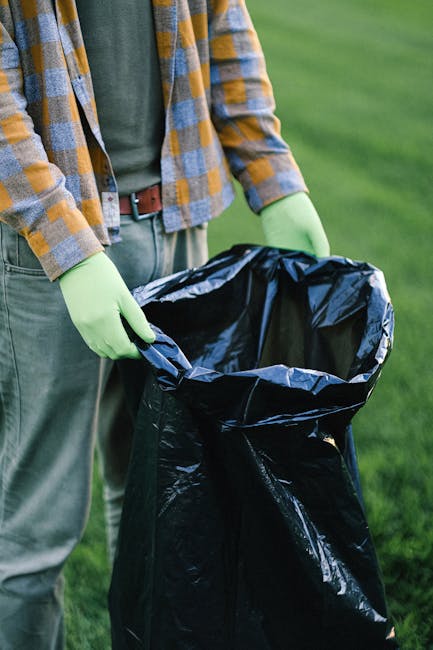 A person standing outdoors on a grassy area, wearing a checkered shirt with shades of yellow, gray, and orange, and light green gloves, is holding open a black plastic rubbish bag. The person's waist